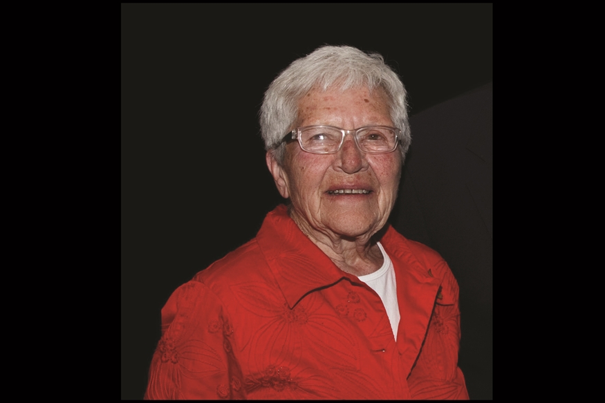 [ai] An elderly woman with short gray hair wearing glasses and a red shirt, smiling while standing in front of a dark background.